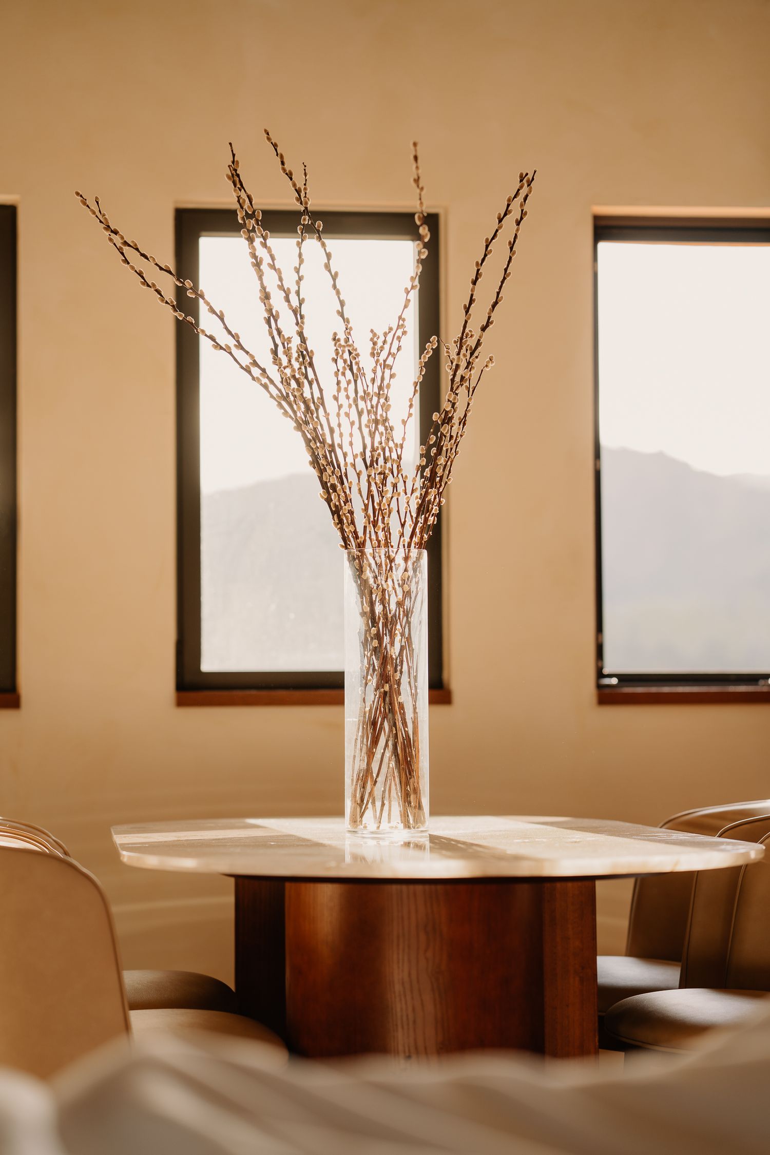 Minimalist sepia-toned panoramic image of twigs in a glass vase on wooden table with windows in background.