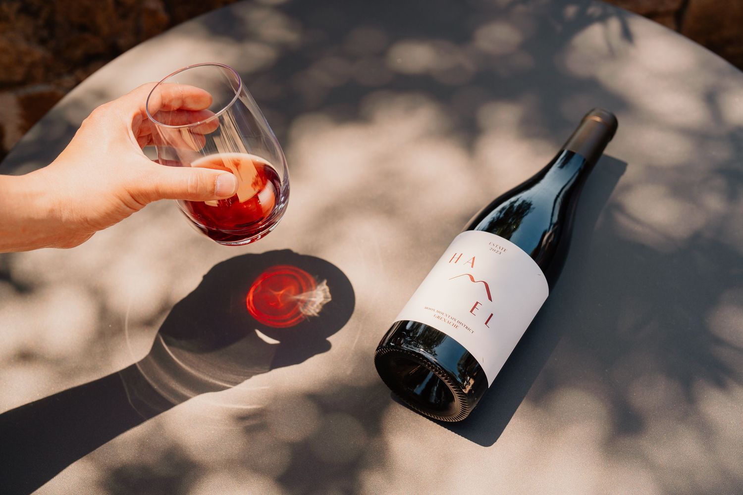 Hand holding red wine glass next to bottle on textured surface with dappled sunlight and shadows.