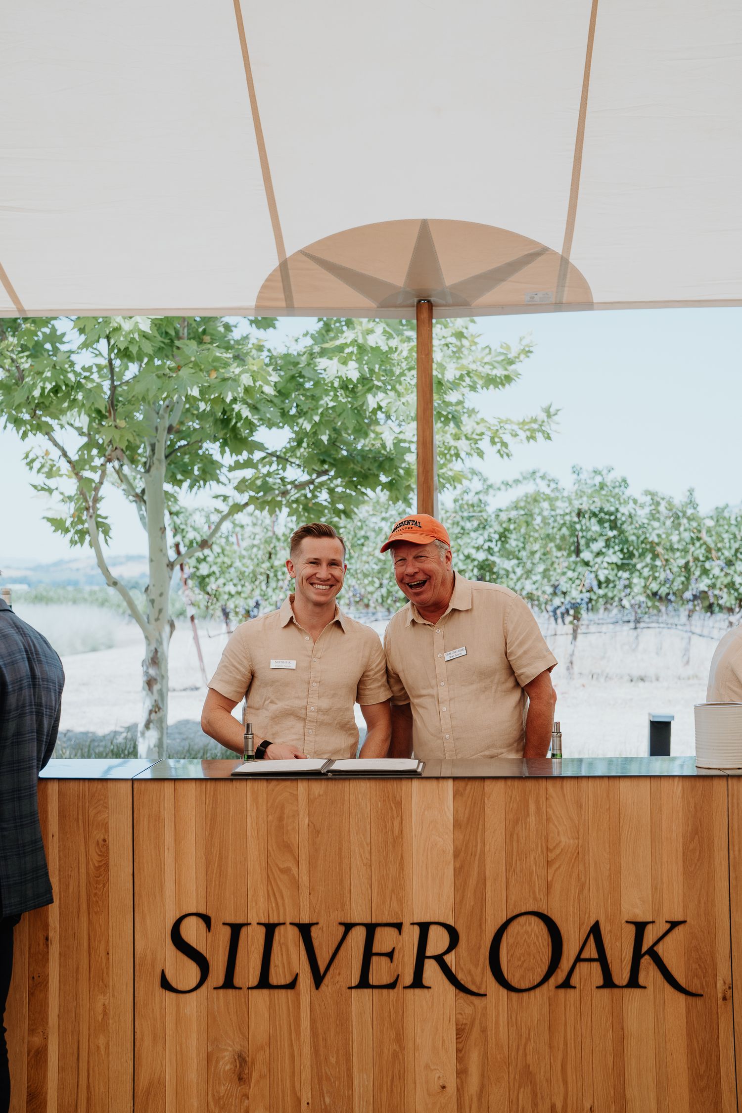 Two smiling Silver Oak winery staff stand behind a wooden welcome reception desk outdoors.