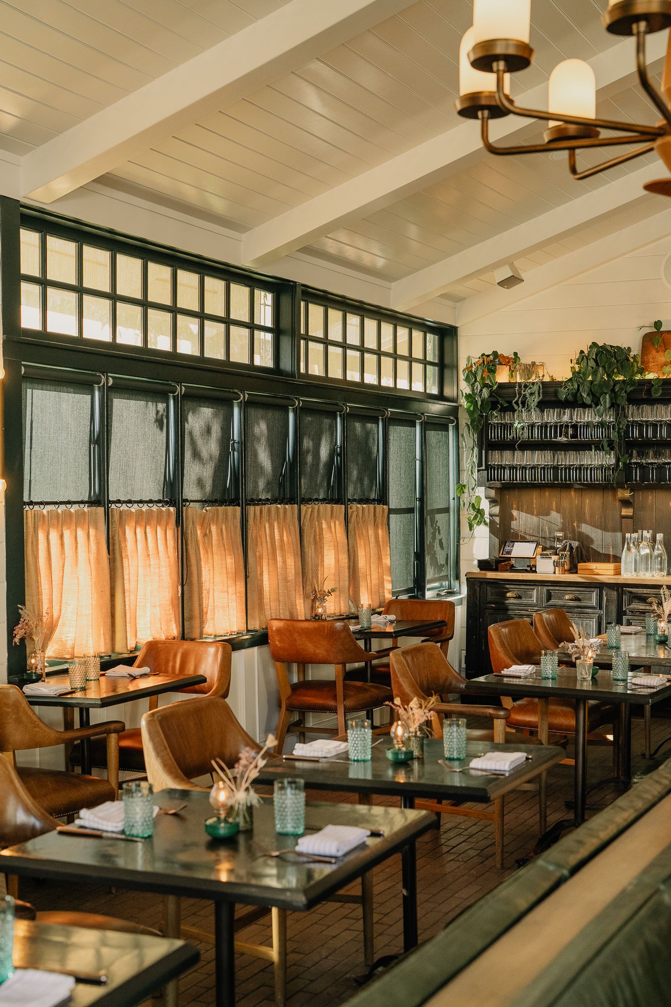 Elegant restaurant interior with leather chairs, set tables, green-framed windows, and warm ambient lighting.