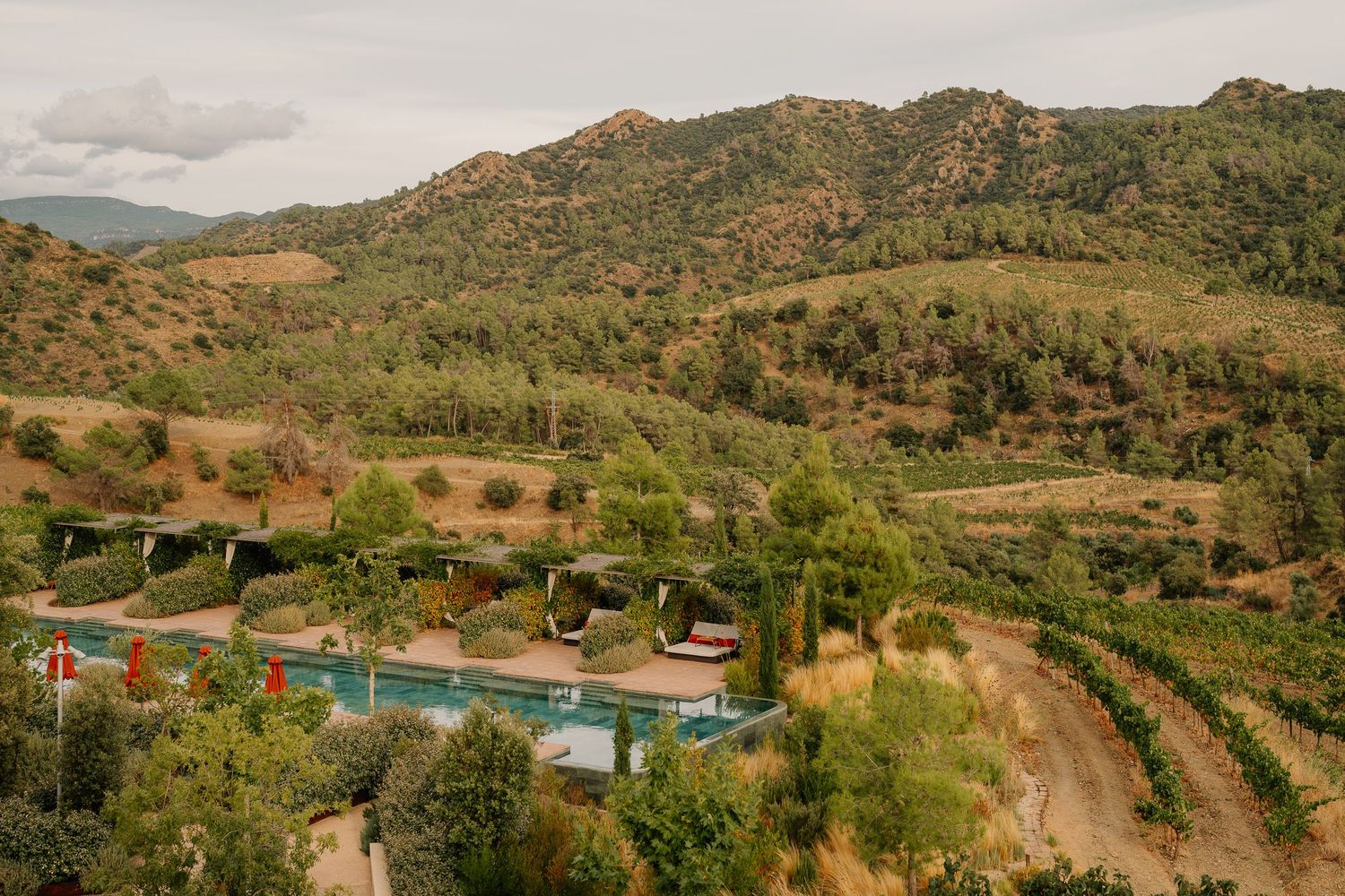Panoramic view of rolling hills with rustic buildings and swimming pools nestled in a lush Mediterranean landscape.