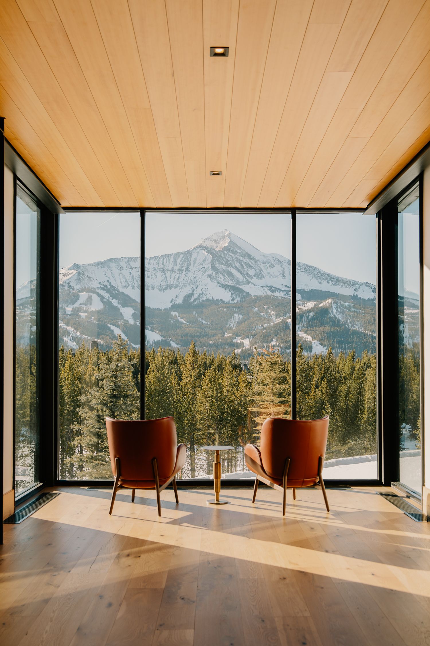 Modern mountain lodge interior with two red chairs facing panoramic windows overlooking a snowy mountain landscape.