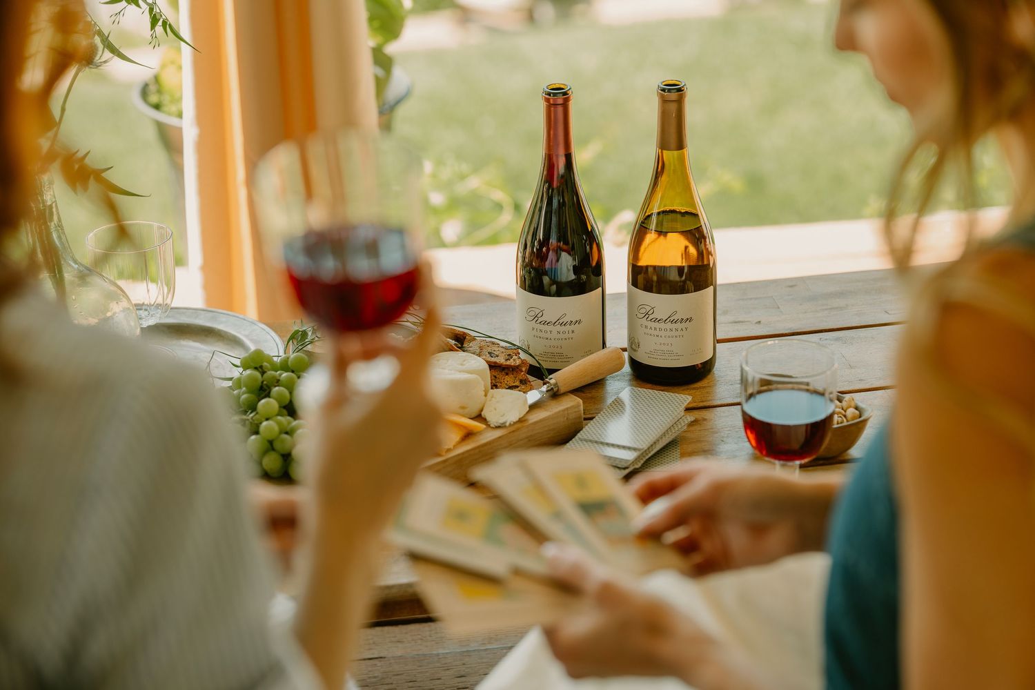 Two women enjoy wine tasting with red and white wine bottles, grapes, and cheese on a rustic wooden table.