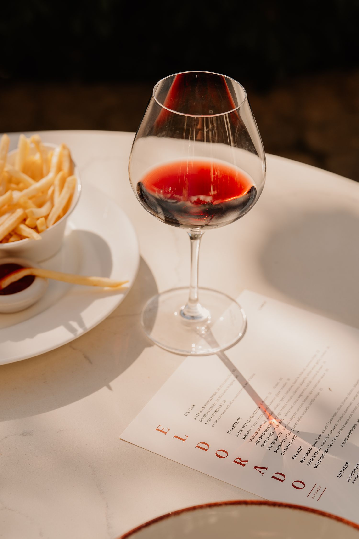 A glass of red wine sits beside a restaurant menu on a white tablecloth with elegant place settings.