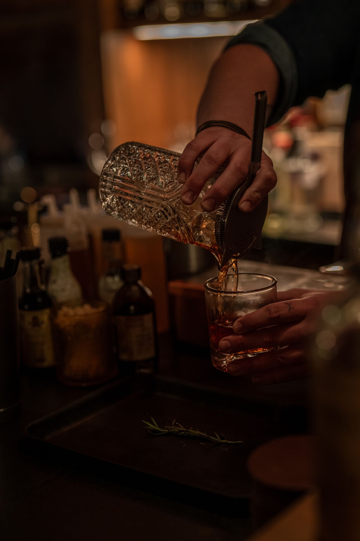 A bartender pours a craft cocktail into a crystal glass in a dimly lit bar setting.