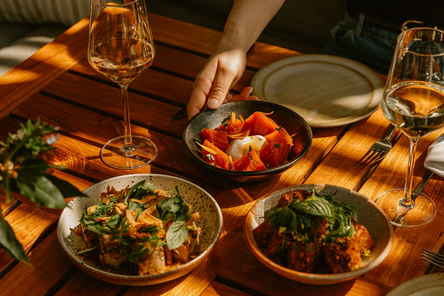 A beautifully set wooden table with colorful dishes, fresh herbs, and wine glasses in warm sunlight.