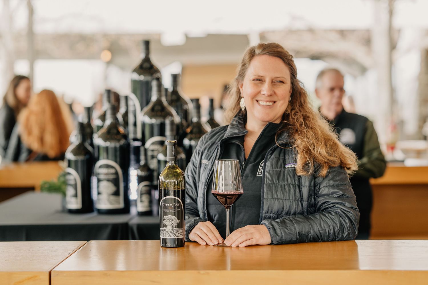Smiling woman poses with Silver Oak wine bottle and glass of red wine at winery tasting counter.