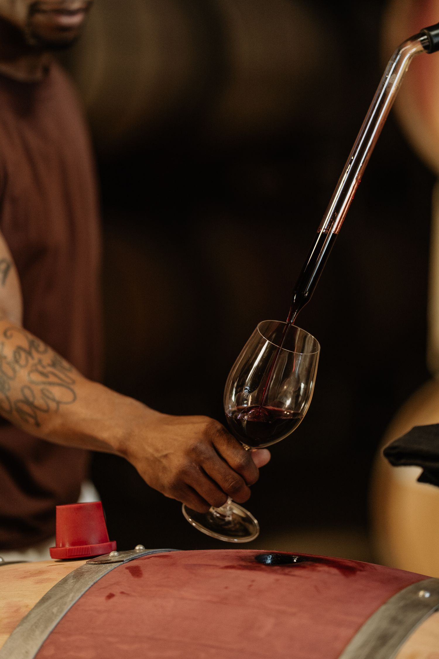 A tattooed winemaker pours red wine from a barrel into a glass at Markham Napa Valley Vineyards.