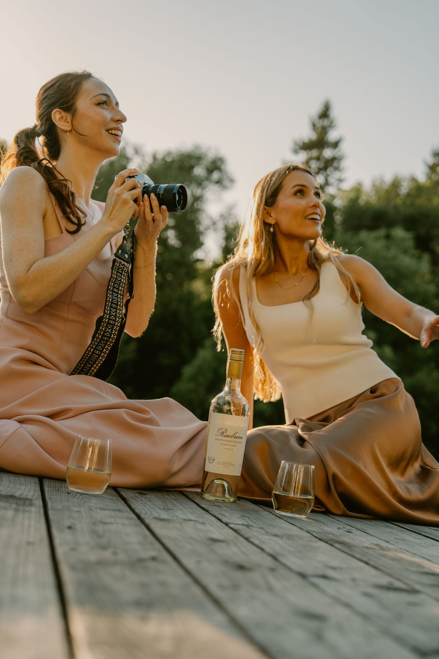 Two women sitting on a wooden dock at sunset, enjoying wine and photography in elegant earth-tone outfits.