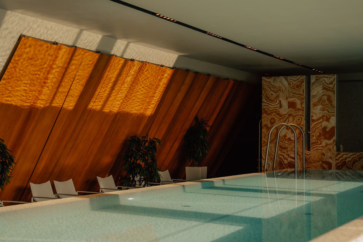 Indoor hotel pool with warm orange drapes, mosaic wall art, and lounge chairs in a serene modern setting.