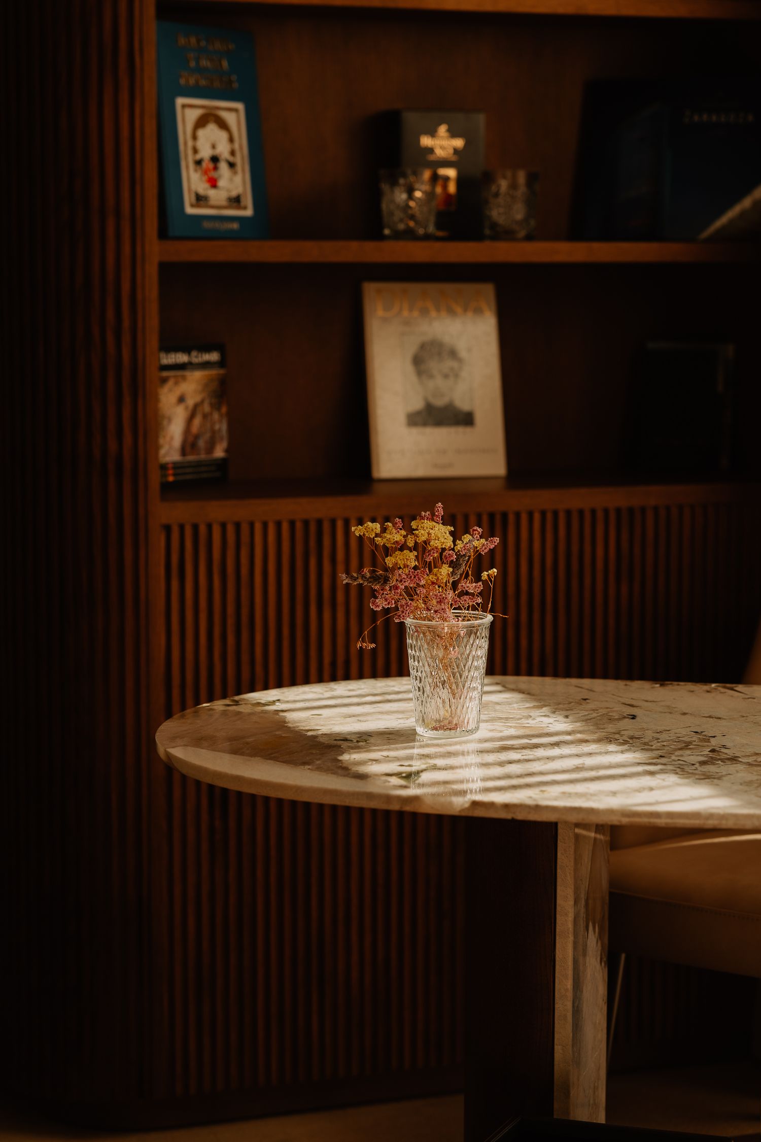 A glass vase with dried flowers sits on a wooden table, casting shadows in warm lighting against wood-paneled walls.