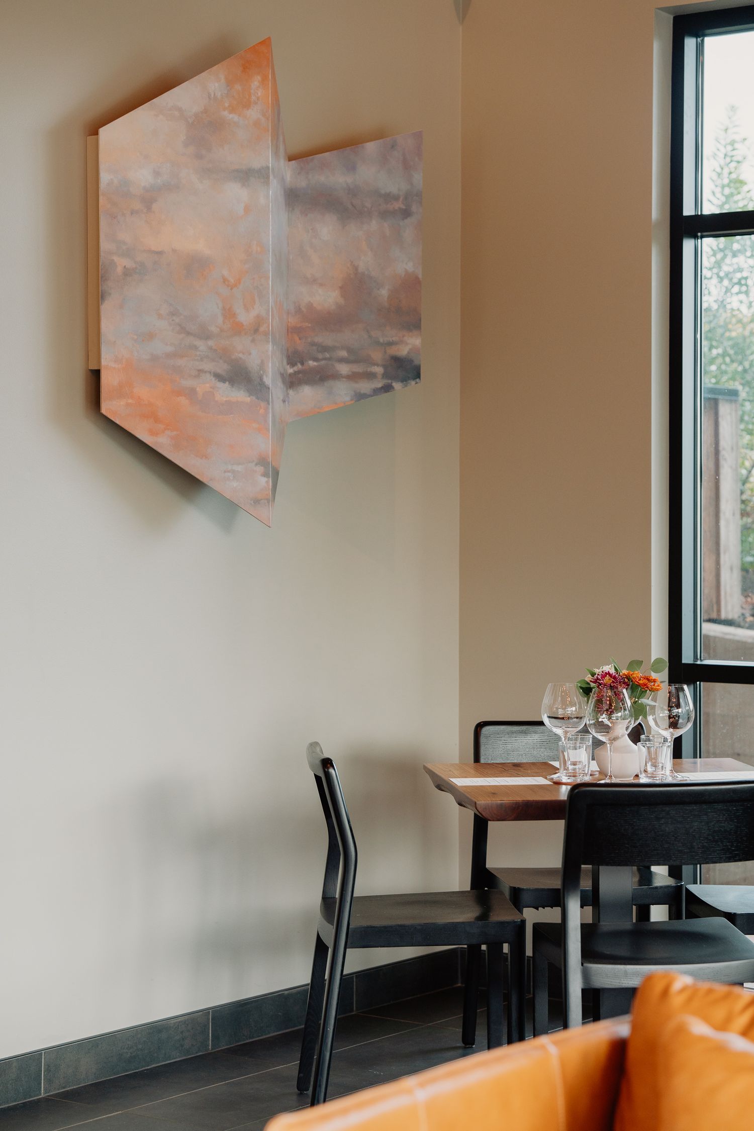 Modern restaurant interior with abstract copper-toned artwork on wall near black dining chairs and window table setting.