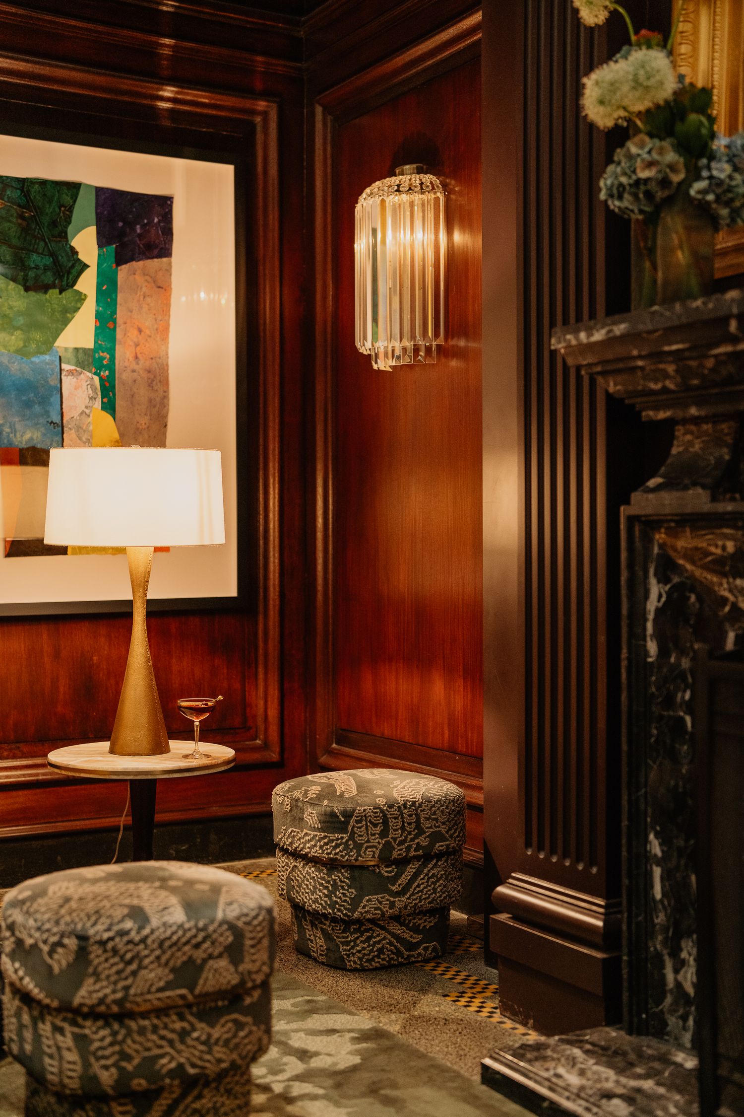 Elegant lounge corner with art-deco style lamp, modern abstract artwork and patterned ottomans against rich wood paneling.