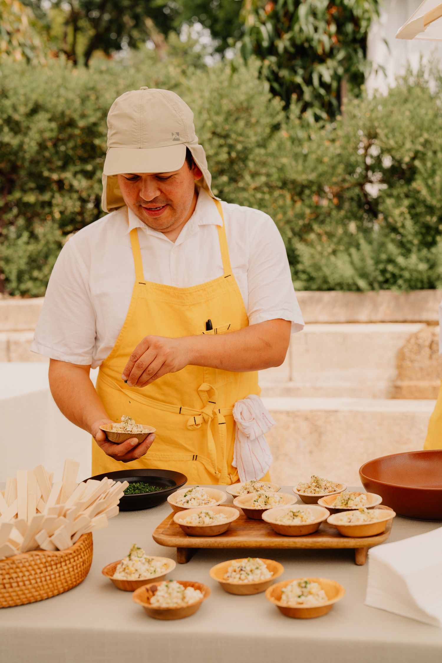 Chef in yellow apron and baseball cap prepares appetizers at an outdoor catering station.