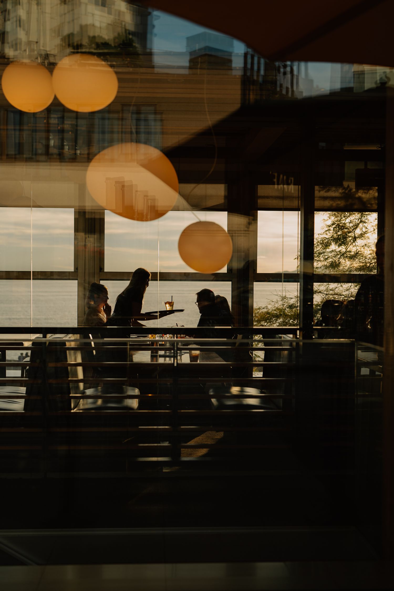 Silhouettes against large windows in a modern interior space with orange globe lights and sunset ambiance.