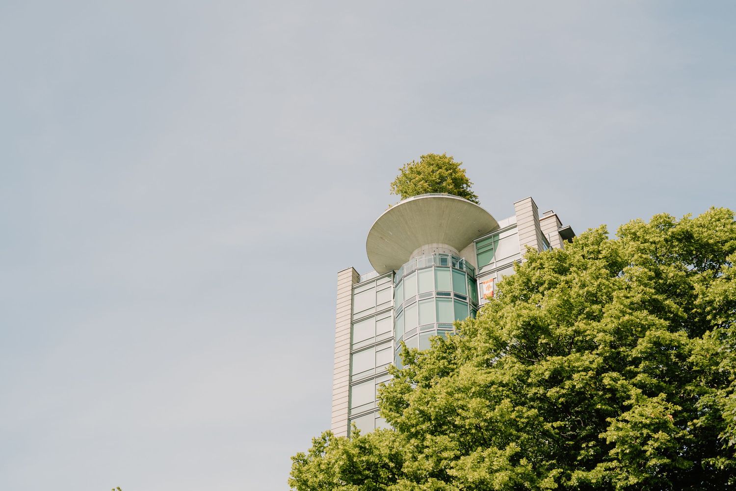 Modern architectural observation tower with glass windows rises above lush green trees against a pale blue sky.