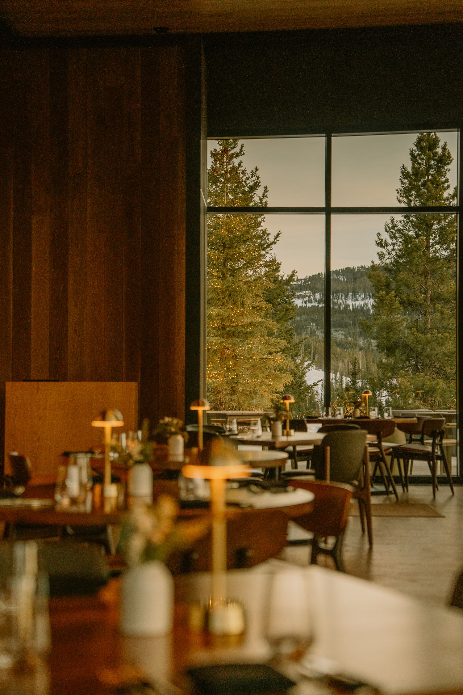 Warm wood-paneled dining room with large windows framing mountain and forest views in golden light.