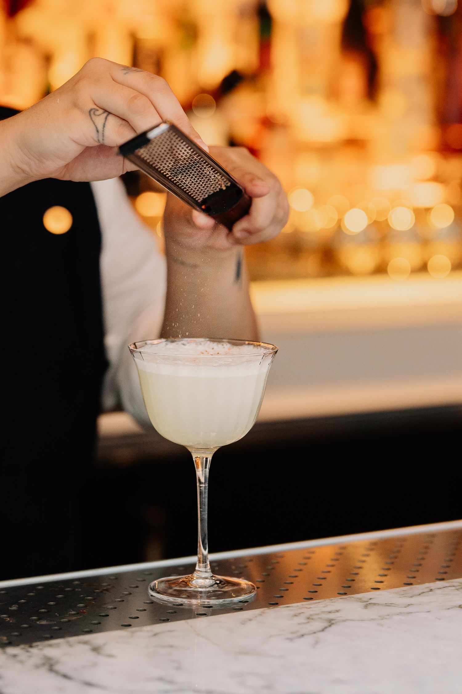 A bartender grates spices over a salt-rimmed cocktail glass with blurred warm lighting in the background.