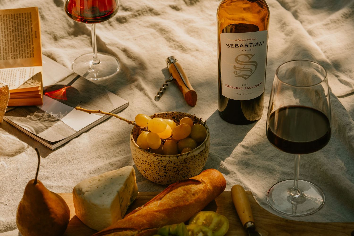 Sebastiani wine bottle with red wine glass, bread, and fruit bowl on white linen picnic spread.