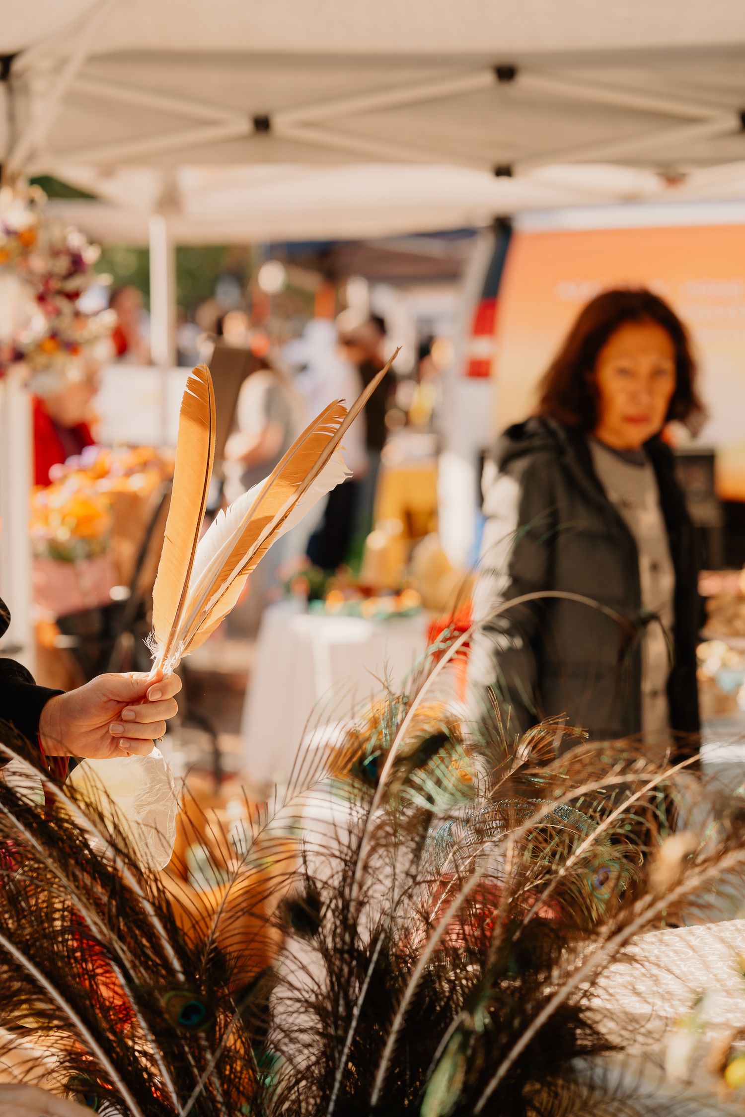 Dried feathers and pampas grass displayed at an outdoor market vendor booth under a white tent canopy.