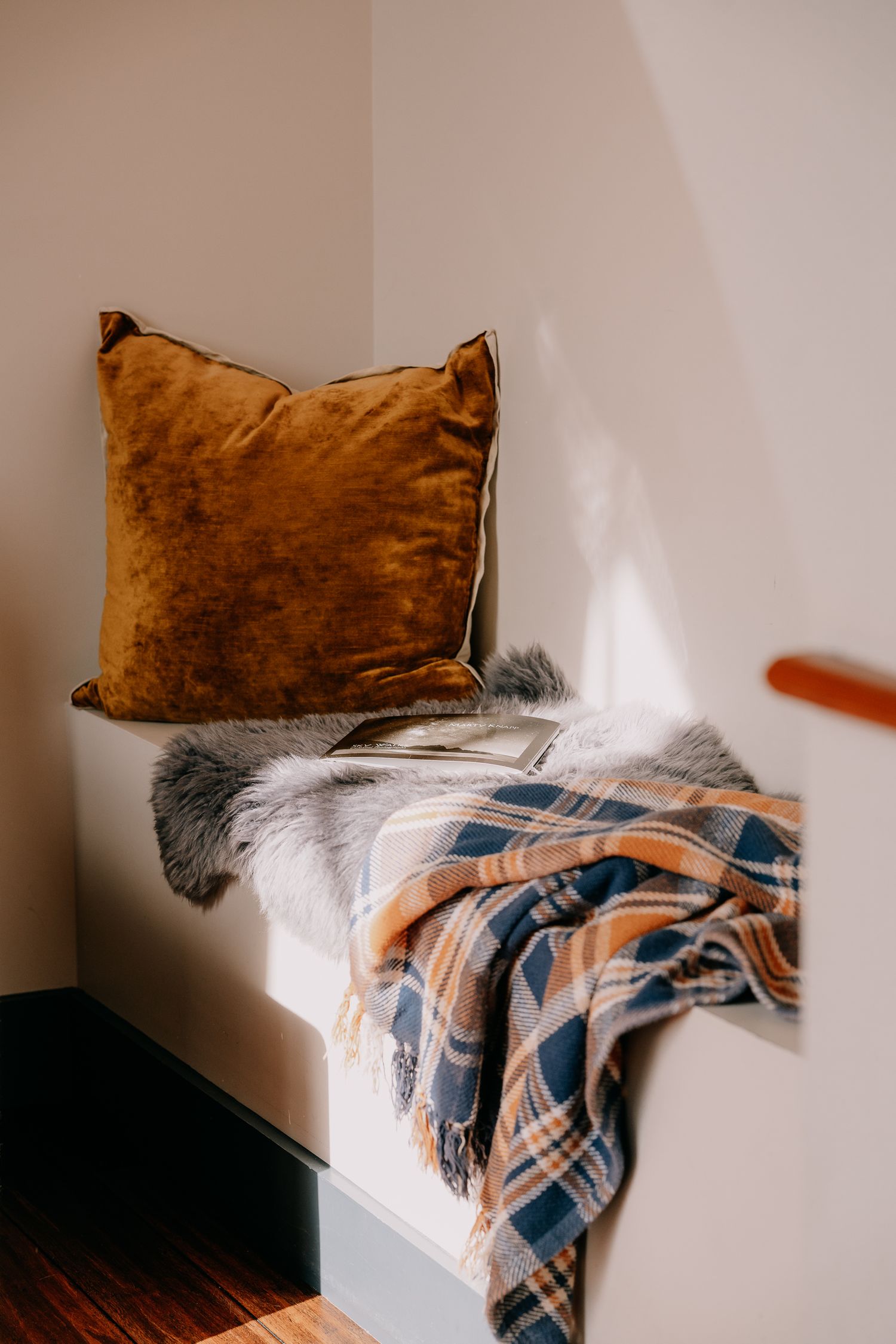 Cozy corner with brown velvet pillow and plaid throw blanket draped over white bench in soft natural lighting.