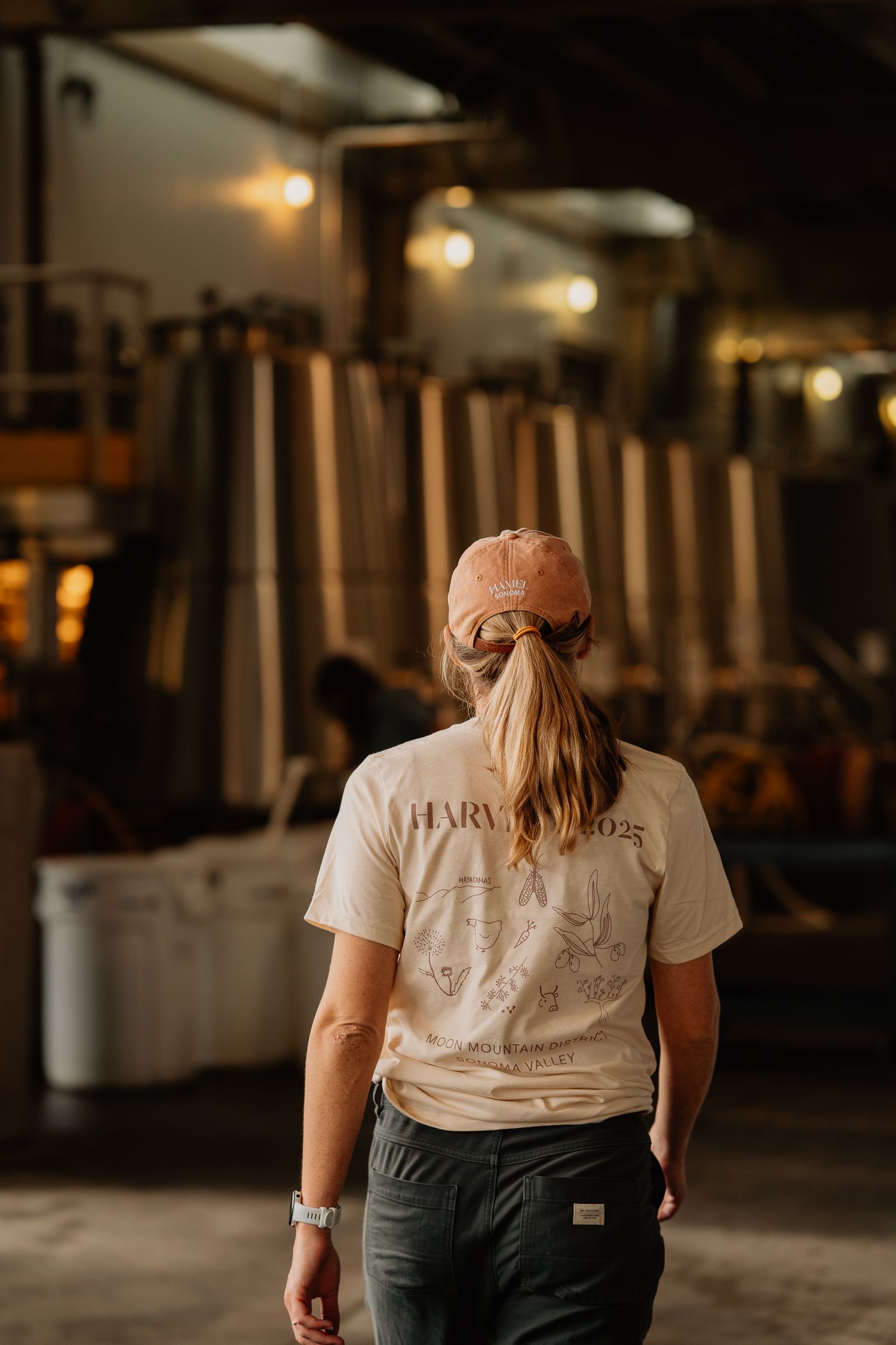 Woman in Harvest 2023 t-shirt and pink cap walks through a dimly lit winery with large fermentation tanks.