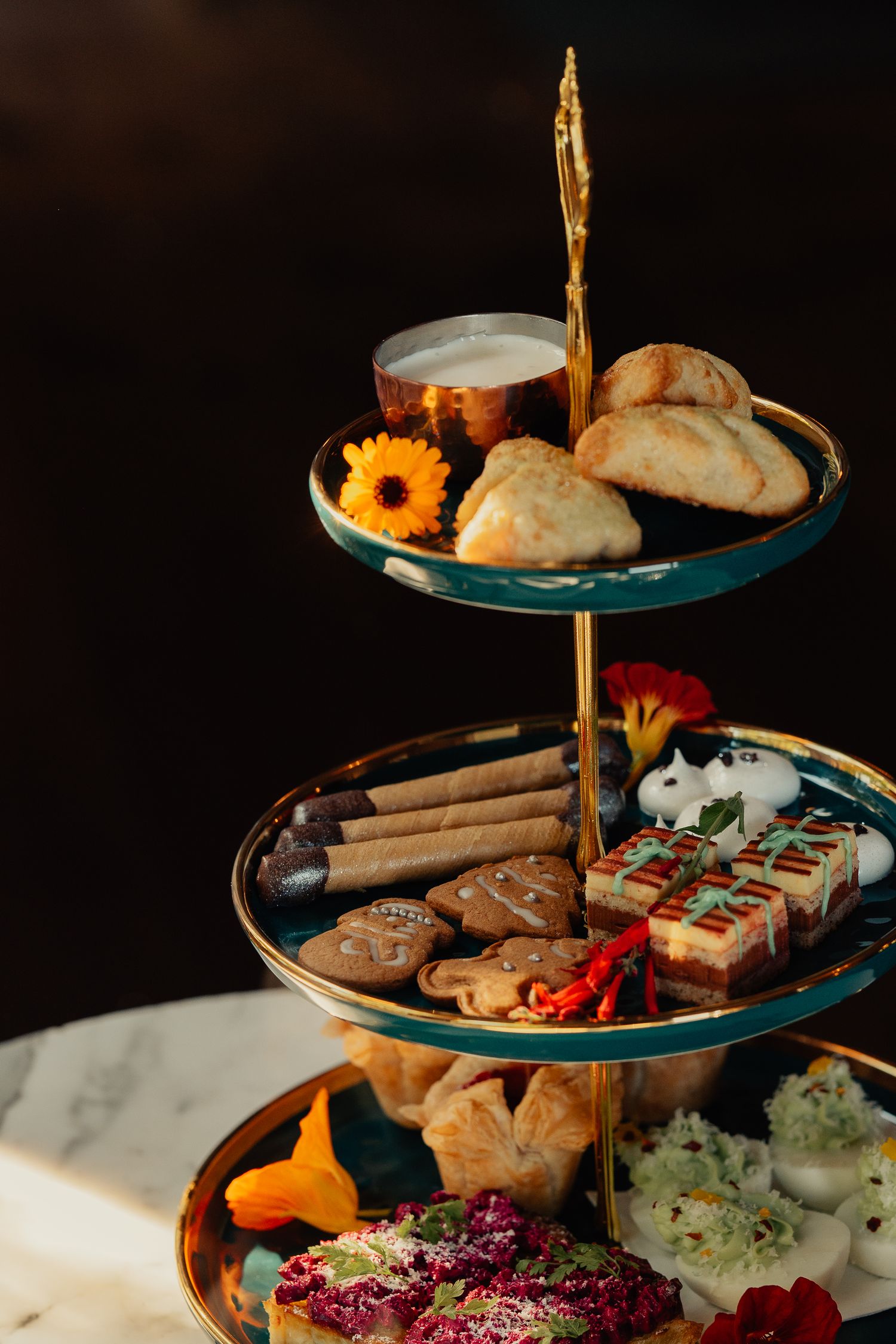 Elegant two-tiered serving platter with pastries, sunflowers, and appetizers on a dark background.