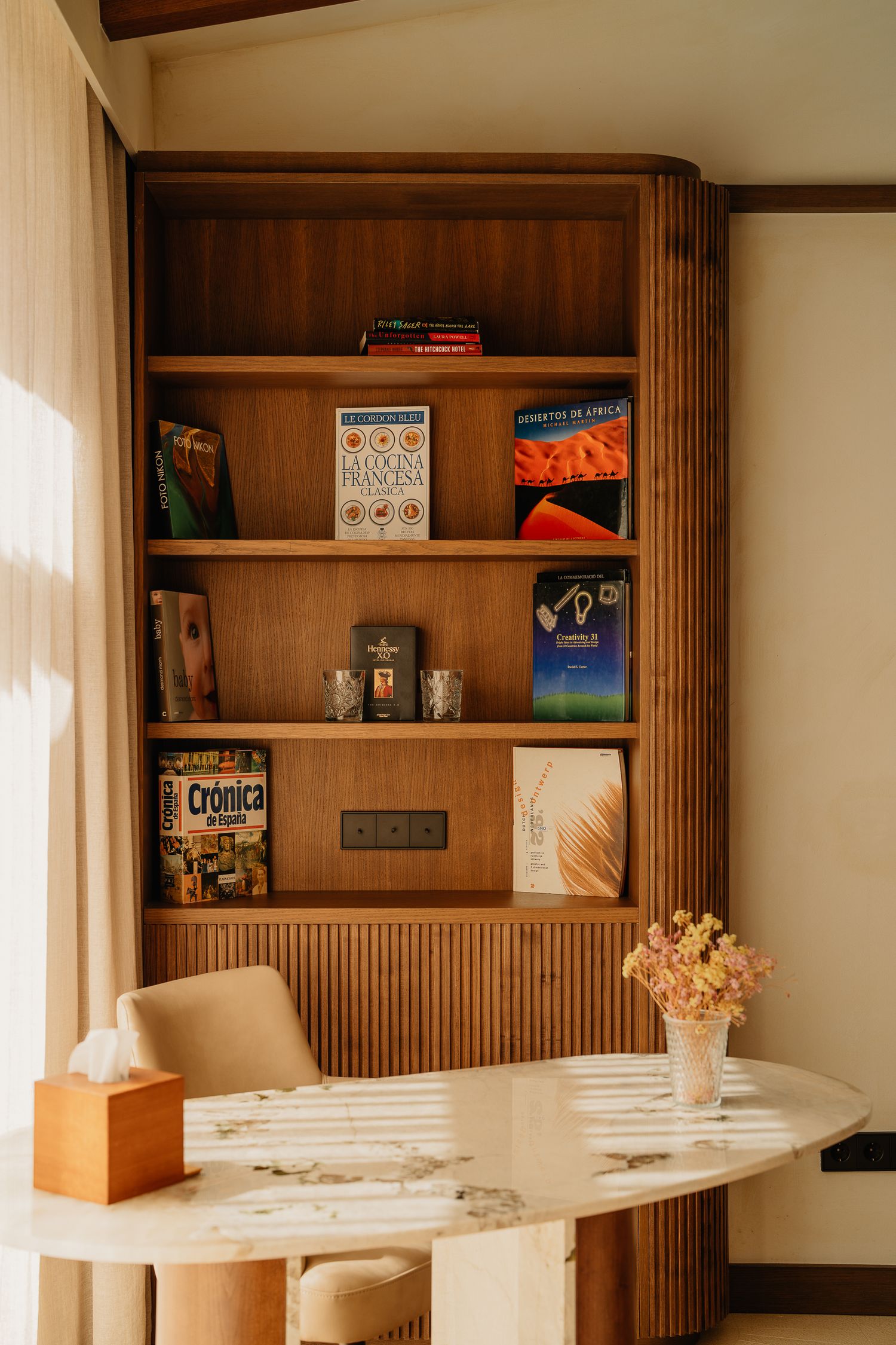 Wooden built-in bookshelf with books and decor next to a round marble table with dried flowers in a vase.