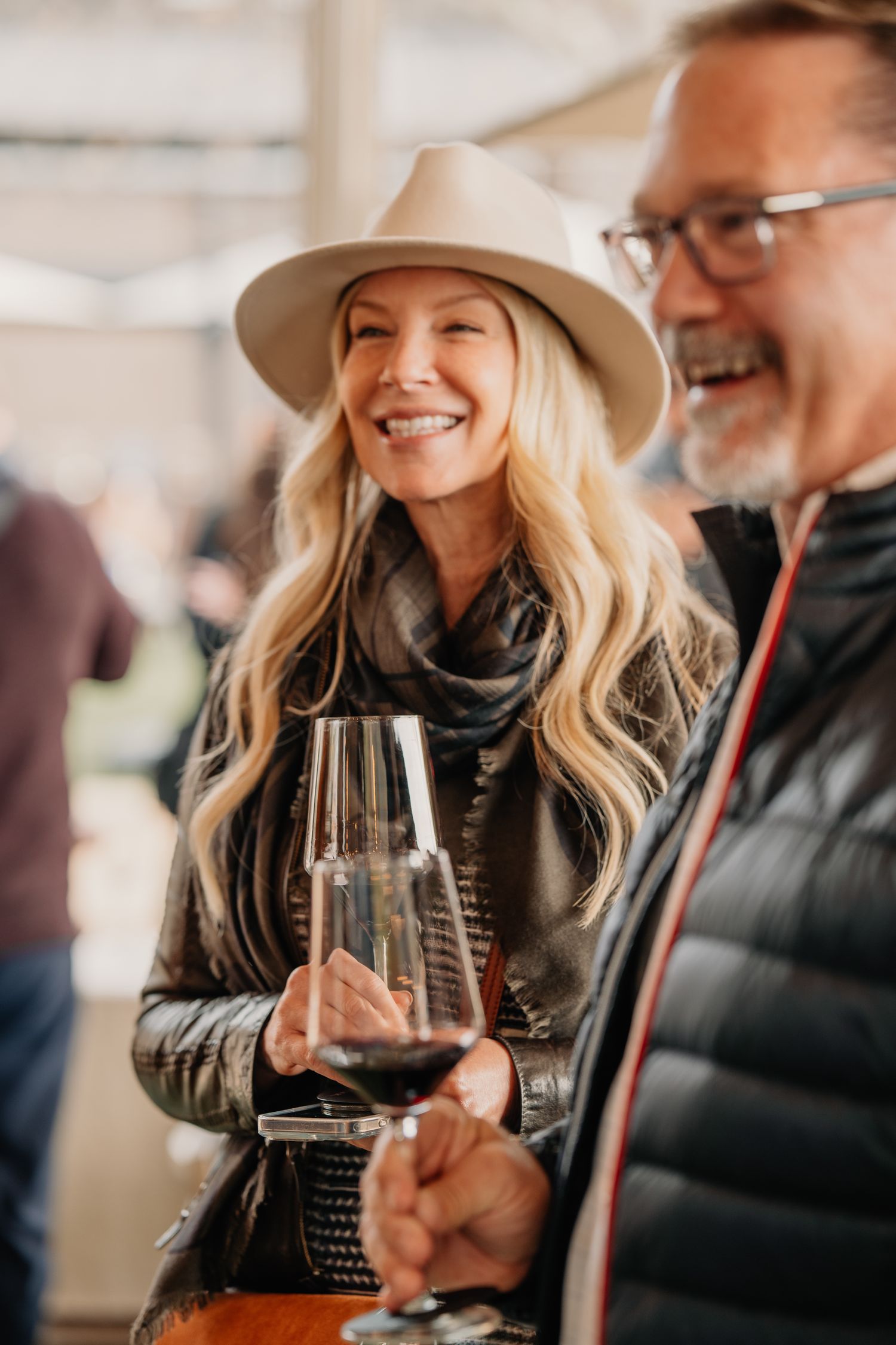 Smiling blonde woman in a wide-brim hat and leather jacket holding a glass of red wine at a winery event.
