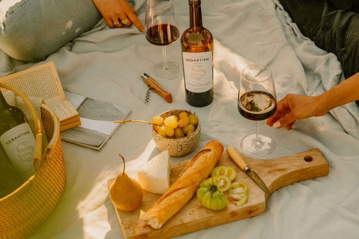 Outdoor picnic spread with Sebastiani wine bottle, charcuterie board, baguette, fruit, and red wine glasses.