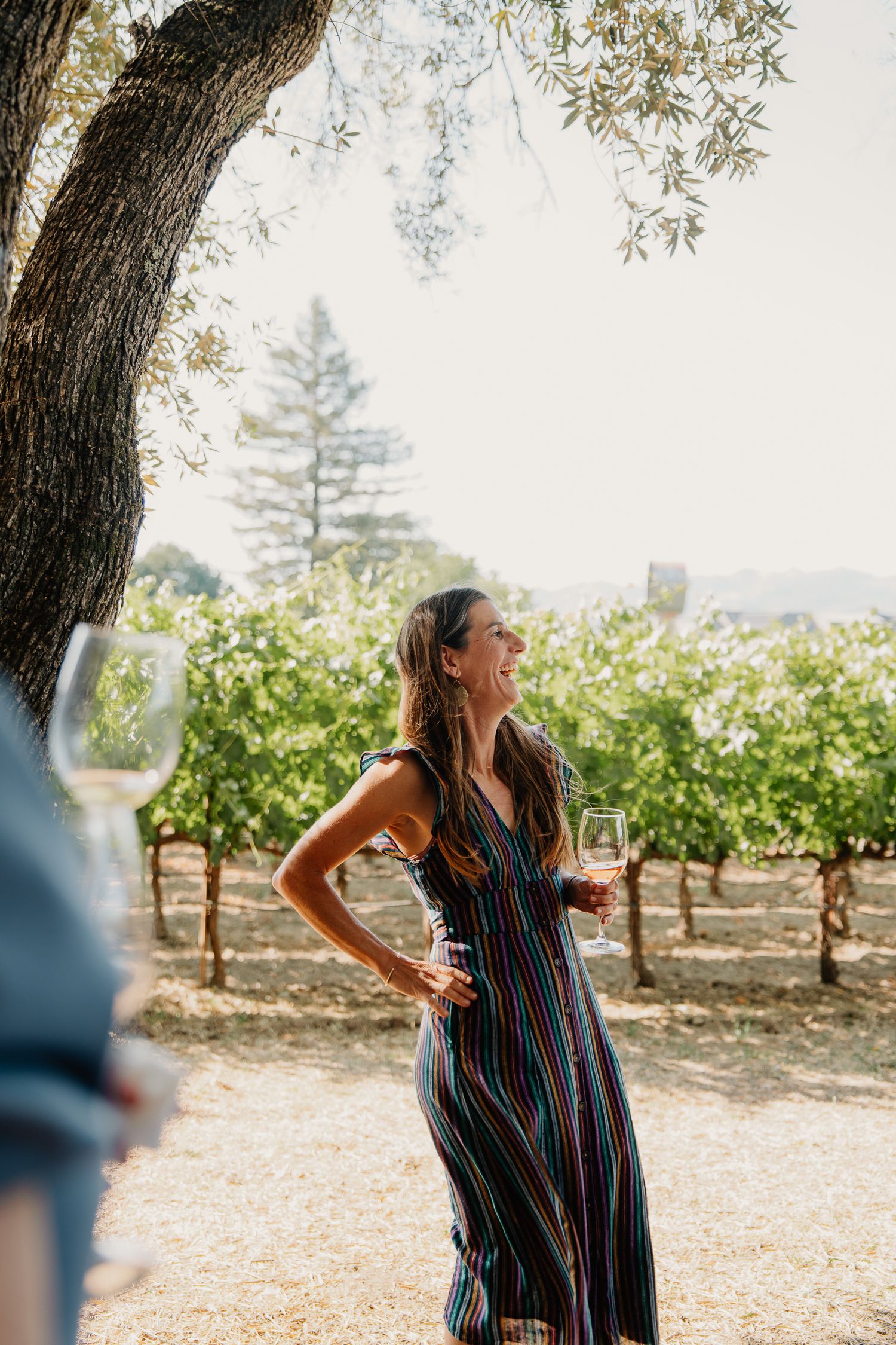Woman in striped dress enjoying wine among vineyard trees on a bright summer day.