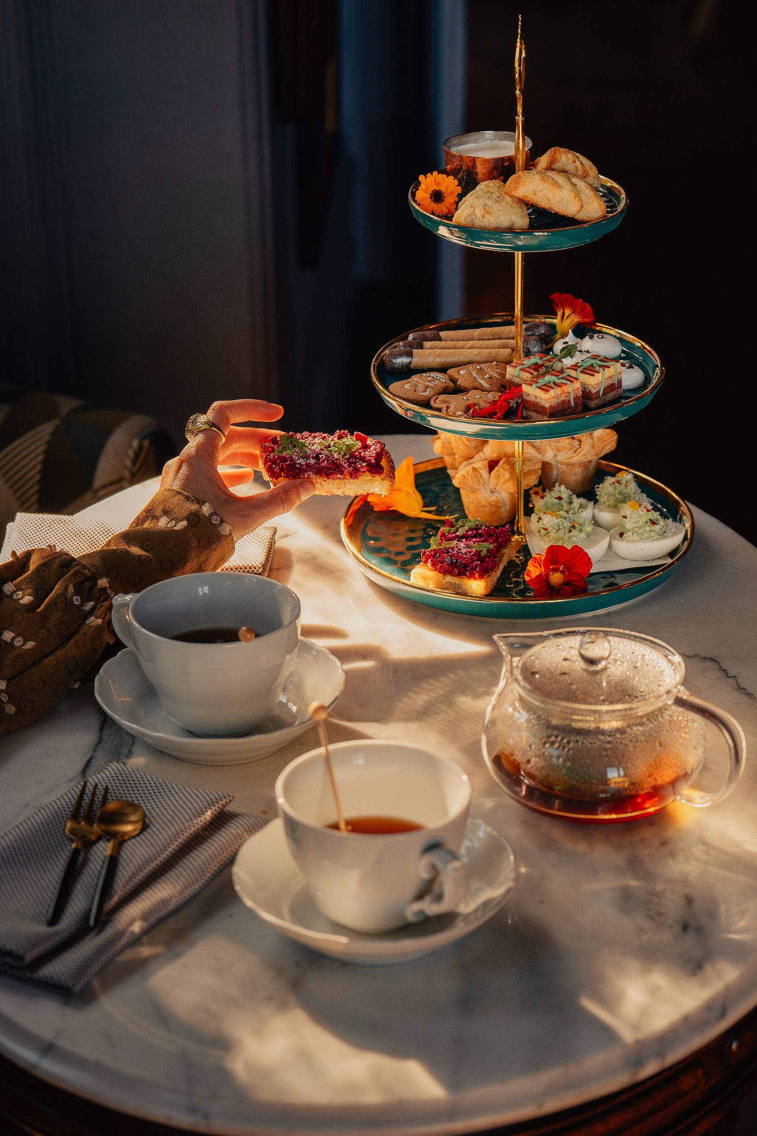 An elegant afternoon tea service with tiered stands displaying pastries, sandwiches and tea cups in natural lighting.