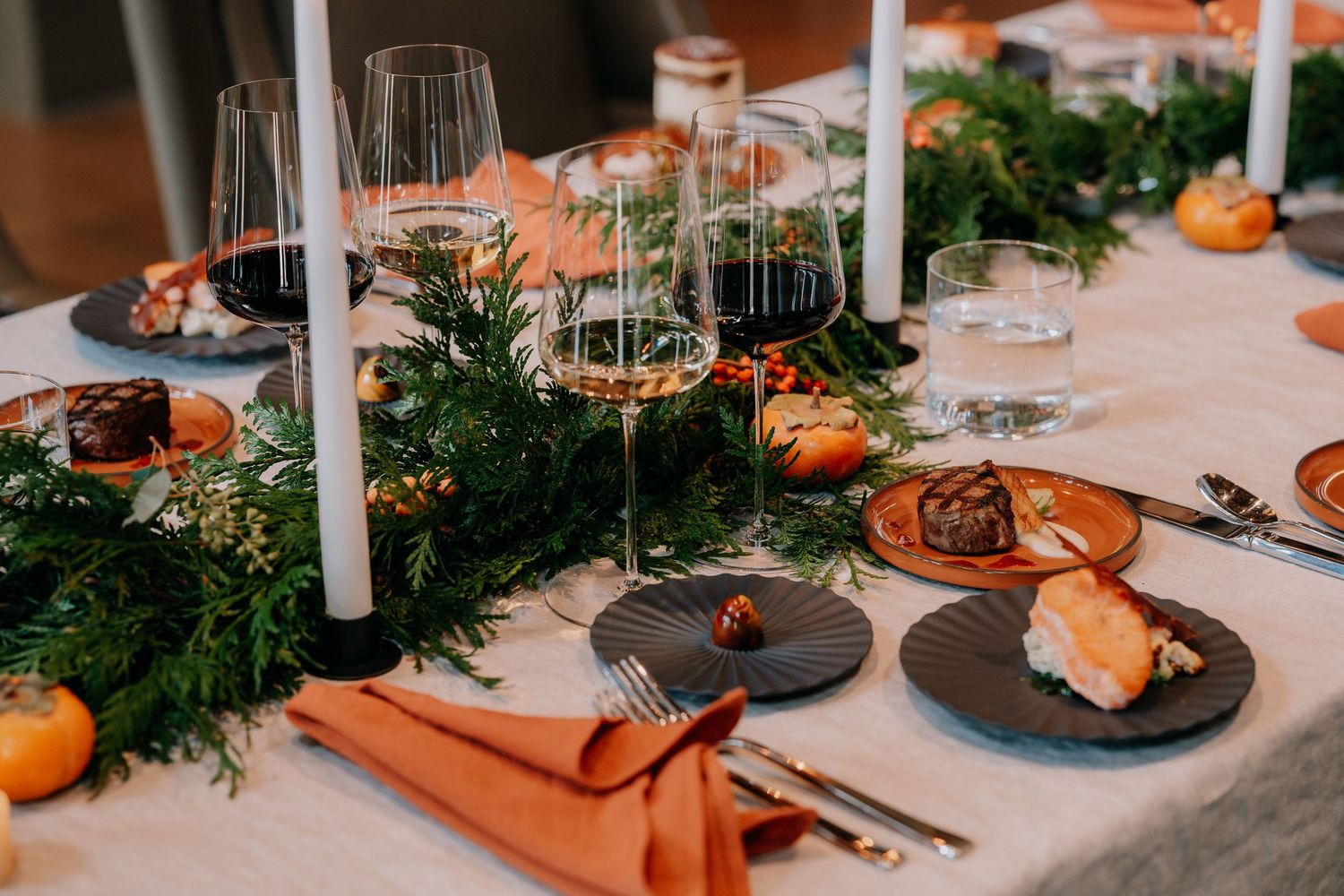 Elegant table setting with wine glasses, greenery, oranges, and white candles on a white tablecloth for holiday dining.