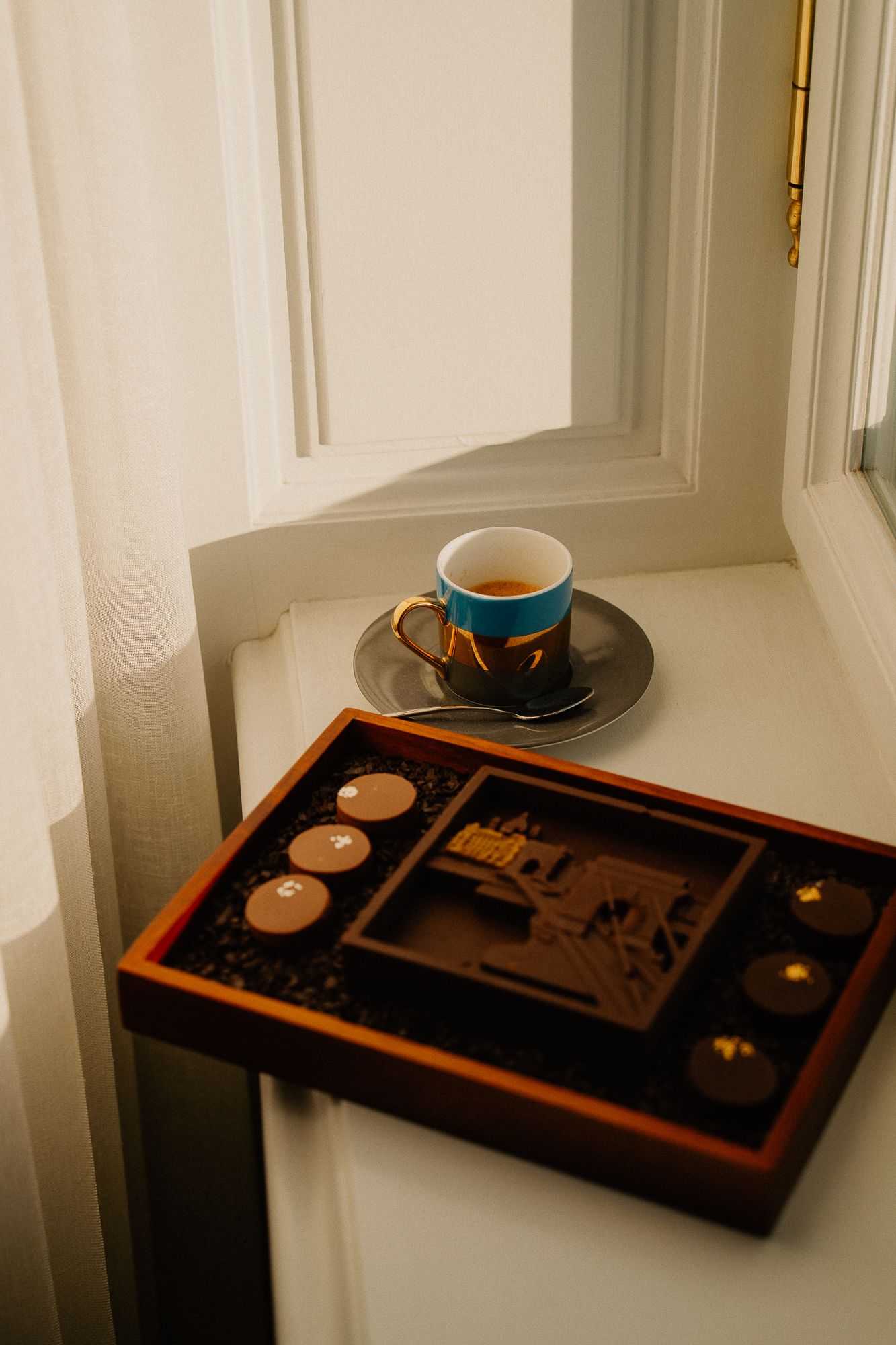 Elegant blue and gold teacup on saucer beside a decorative wooden chocolate box on a sunlit windowsill.