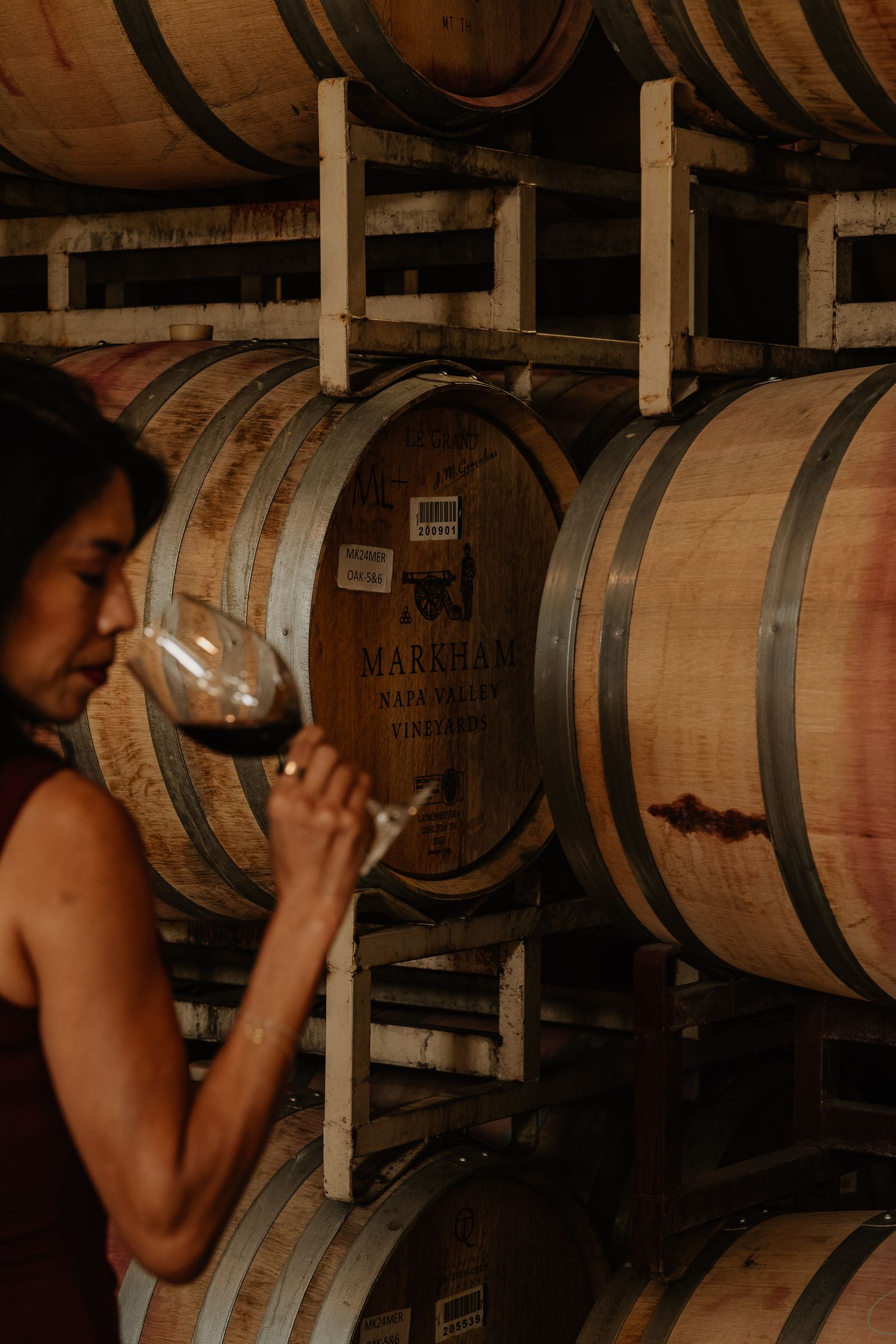 Woman tasting red wine from a glass in front of oak barrels at Markham Napa Valley winery.