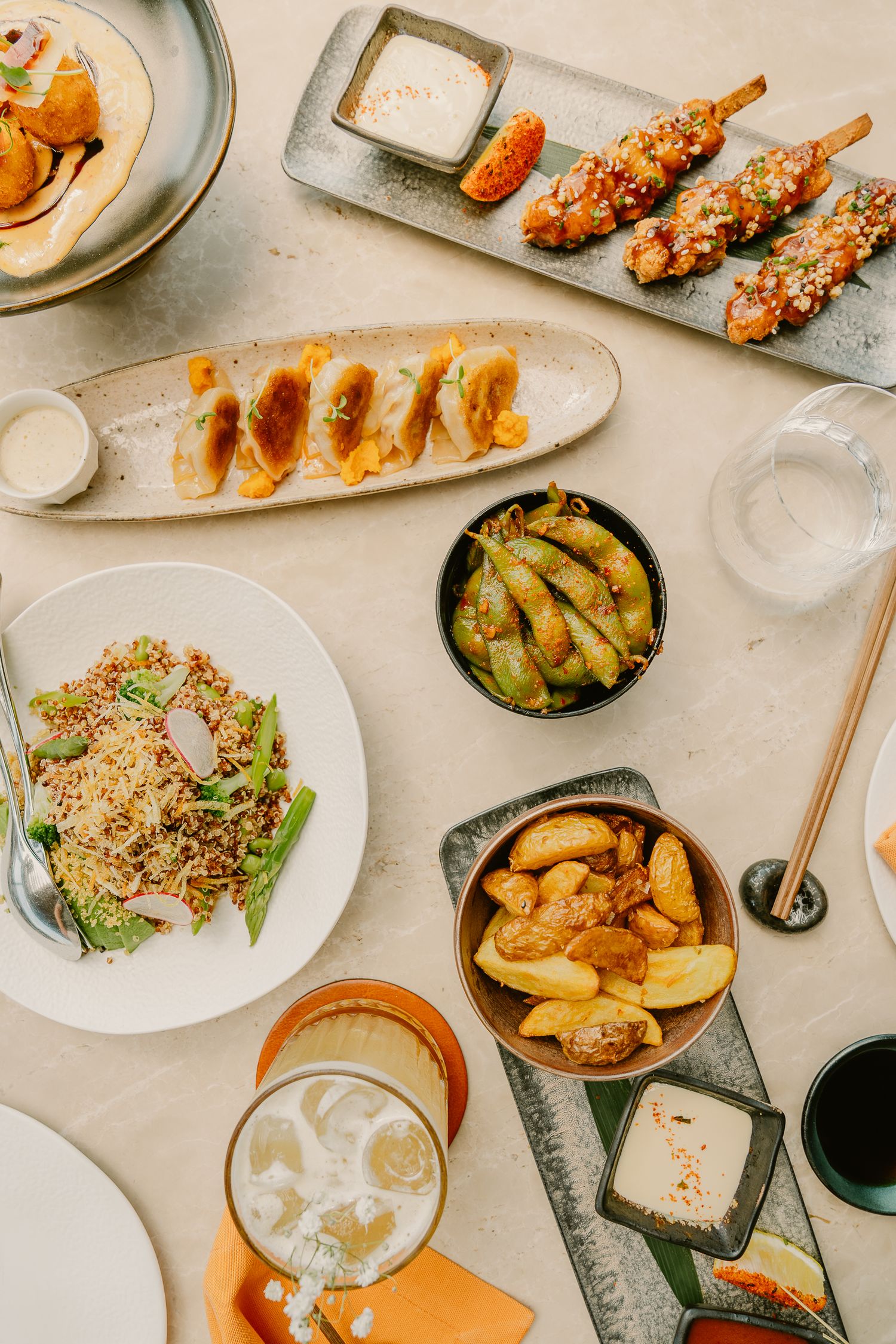 A spread of Asian dishes including dumplings, skewers, edamame, fried potatoes, grain salad, and a beer on a table.