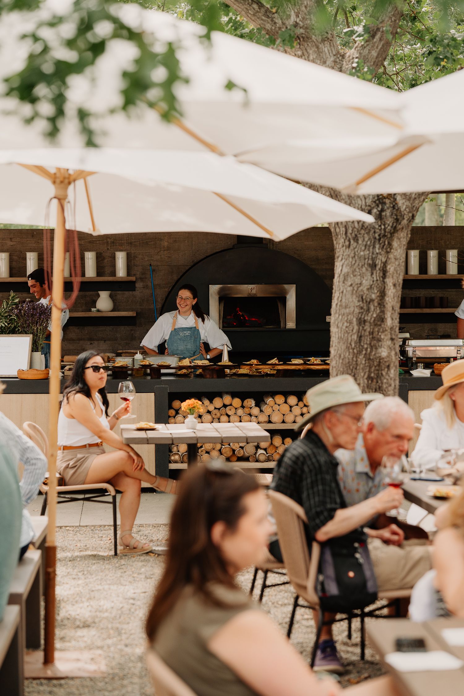 Outdoor cafe with white sun shades and wooden benches showing people dining and a bread display under trees.