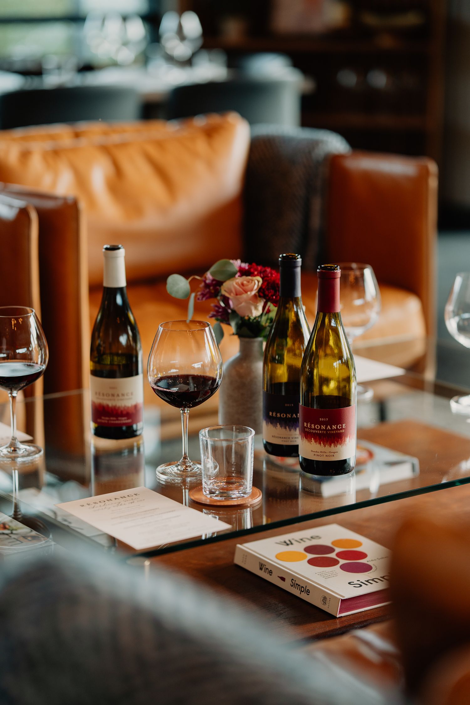 Wine bottles and glasses arranged on a table with caramel leather banquette seating and a small floral centerpiece.