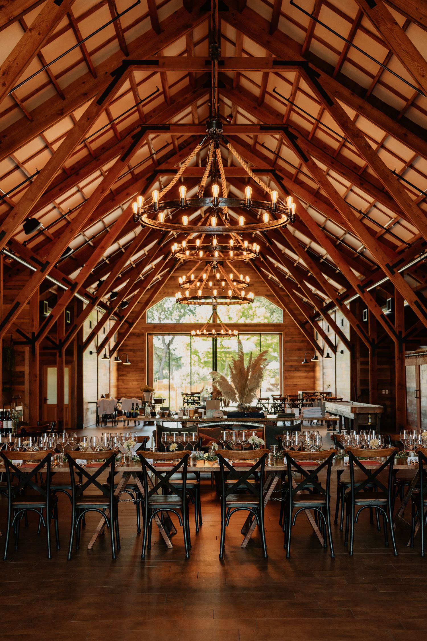 Interior of rustic barn venue with exposed wooden beams, warm lighting from round chandeliers, and dark seating.