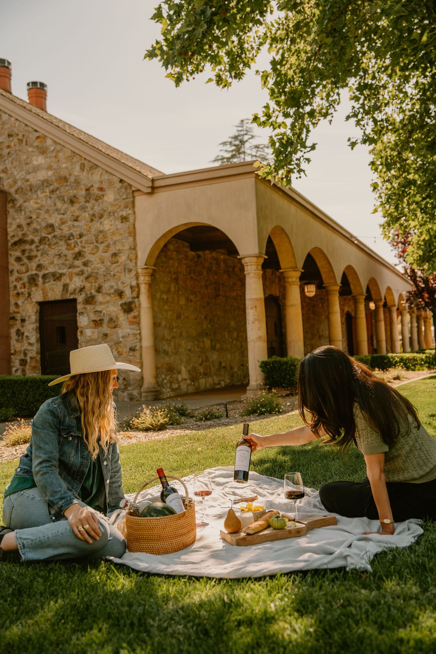 Two women enjoying a wine picnic on a blanket in front of a rustic stone building with arches.