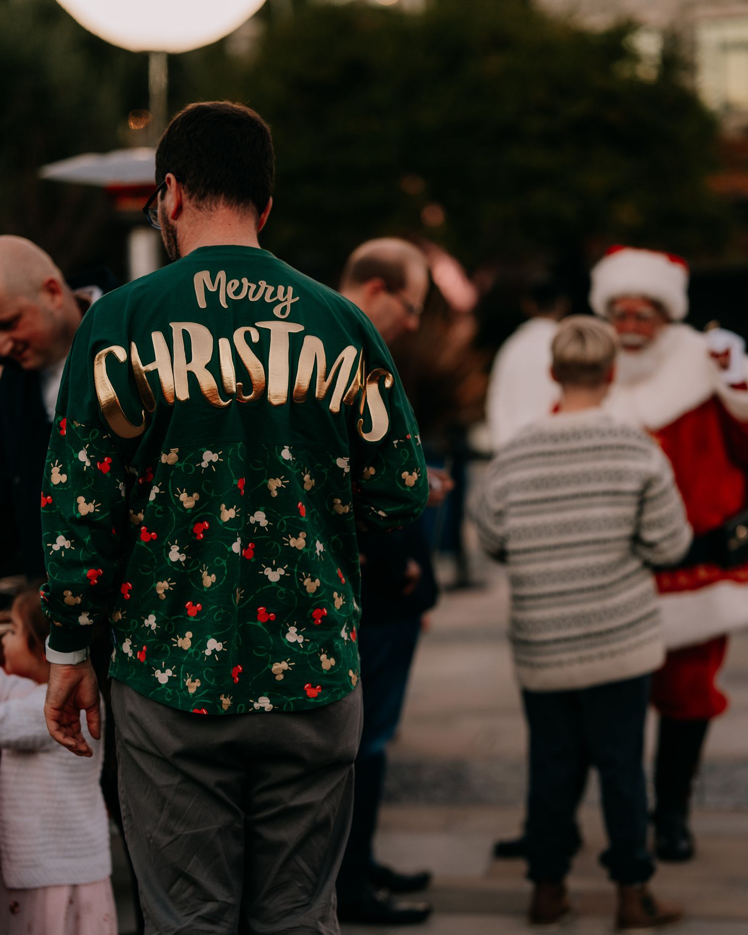 Person wearing a festive green Christmas sweater with reindeer pattern stands in a crowd near someone dressed as Santa.
