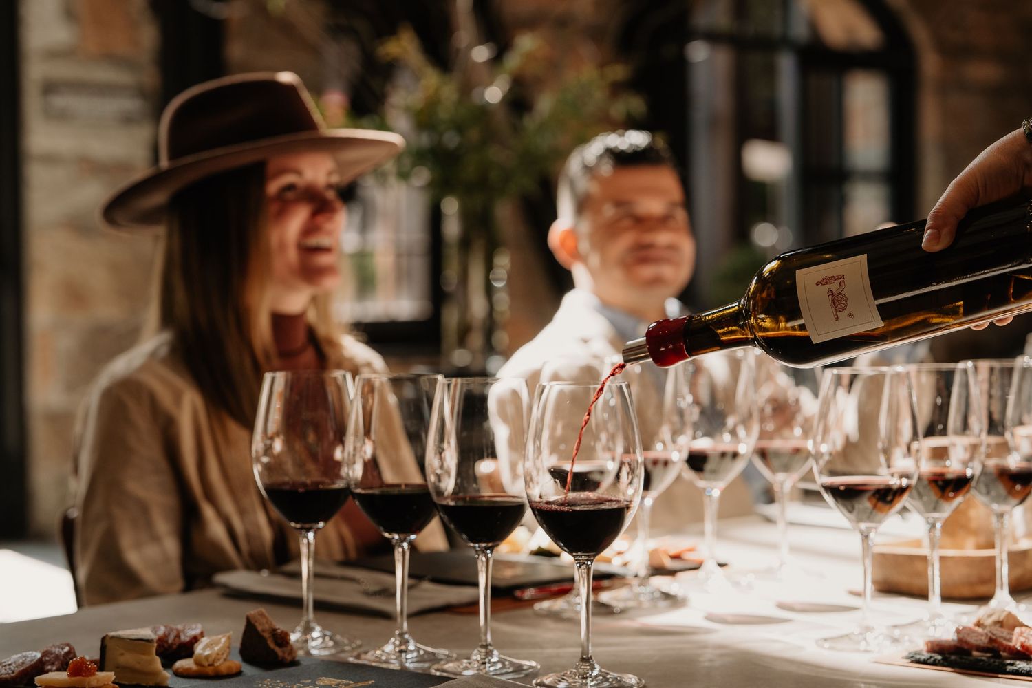 Red wine being poured into glasses during an elegant wine tasting event with guests seated at a rustic table.