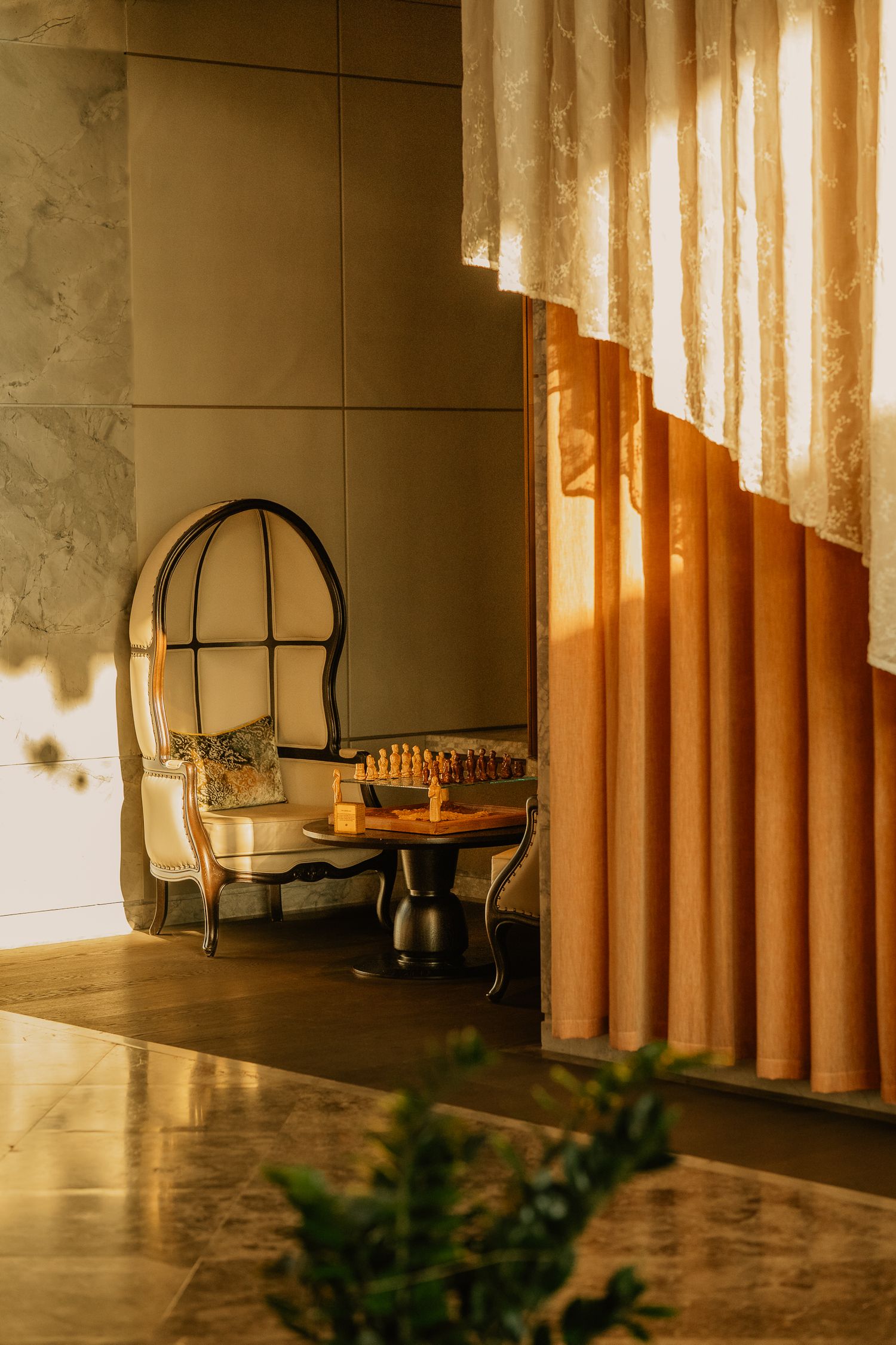 Cozy nook with vintage arched chair, chess board table, and golden curtains bathed in warm sunset light.