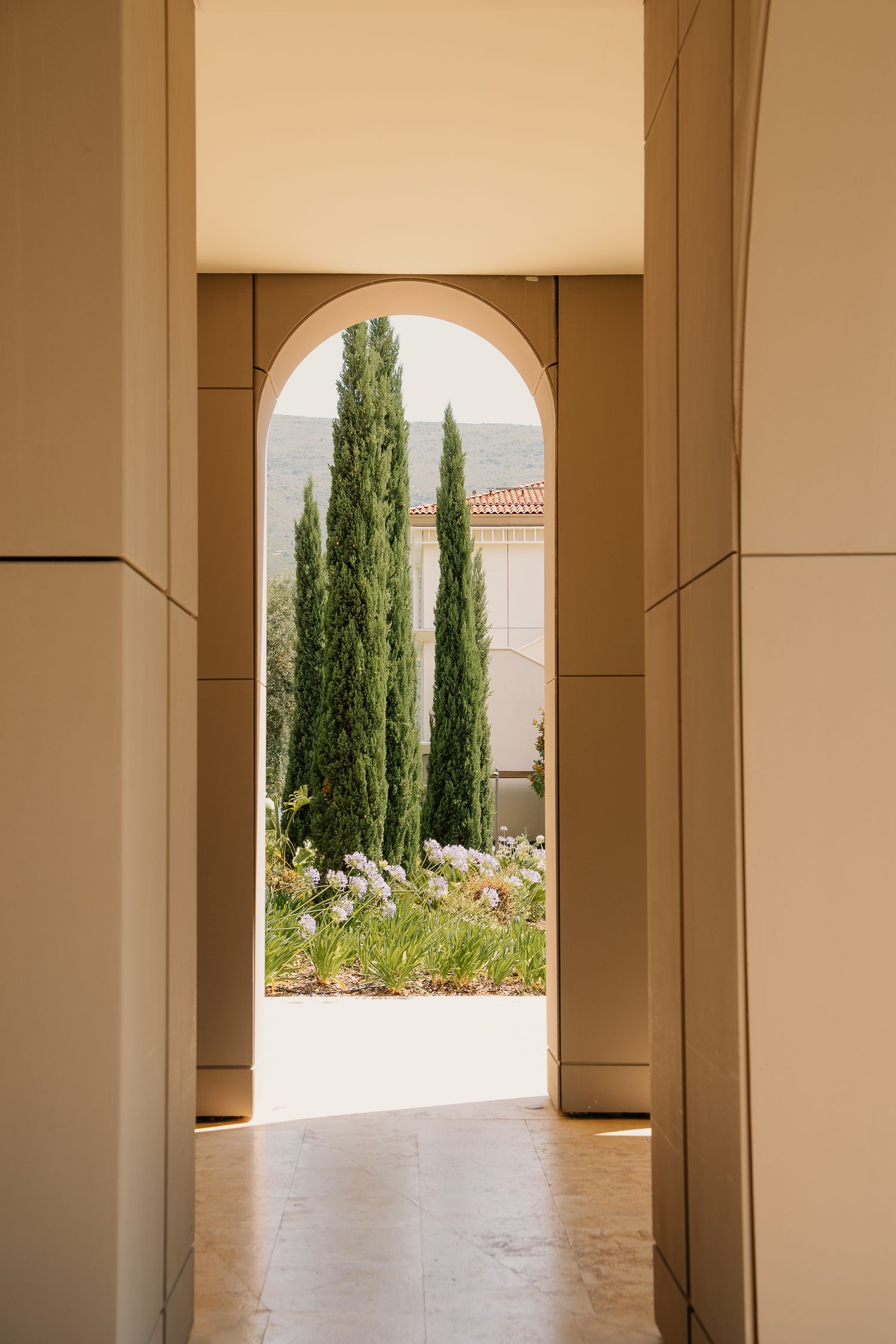Elegant beige hallway with arched doorway framing tall cypress trees and a Mediterranean-style garden beyond.