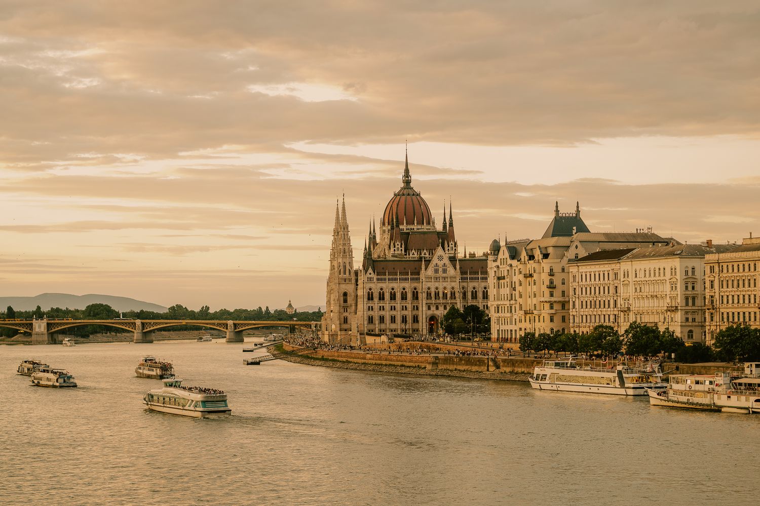 Budapest Parliament Building along the Danube River at golden sunset with boats passing under Margaret Bridge.
