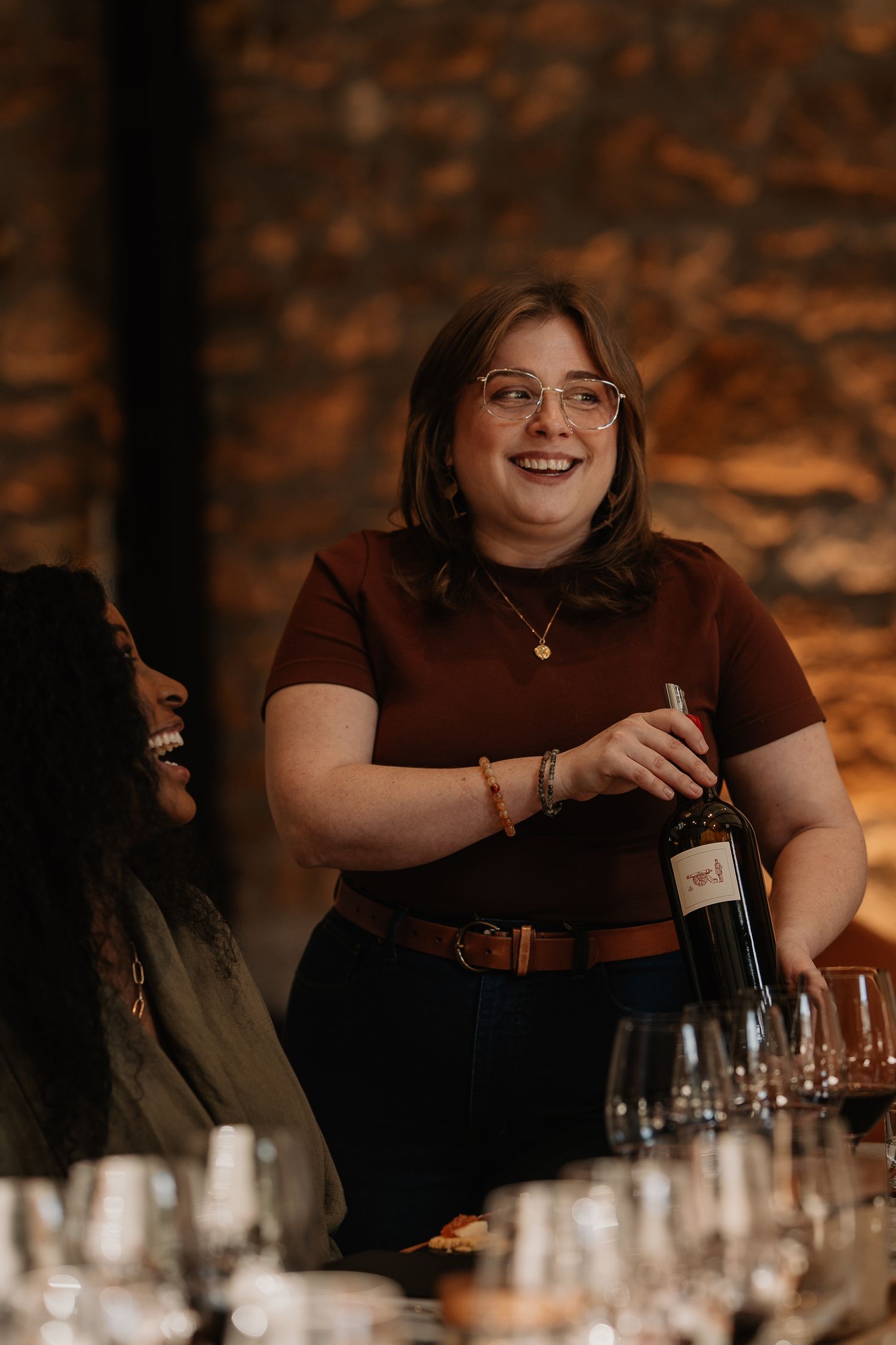 Cheerful woman in glasses presenting a bottle of wine at a rustic stone cellar wine tasting event.