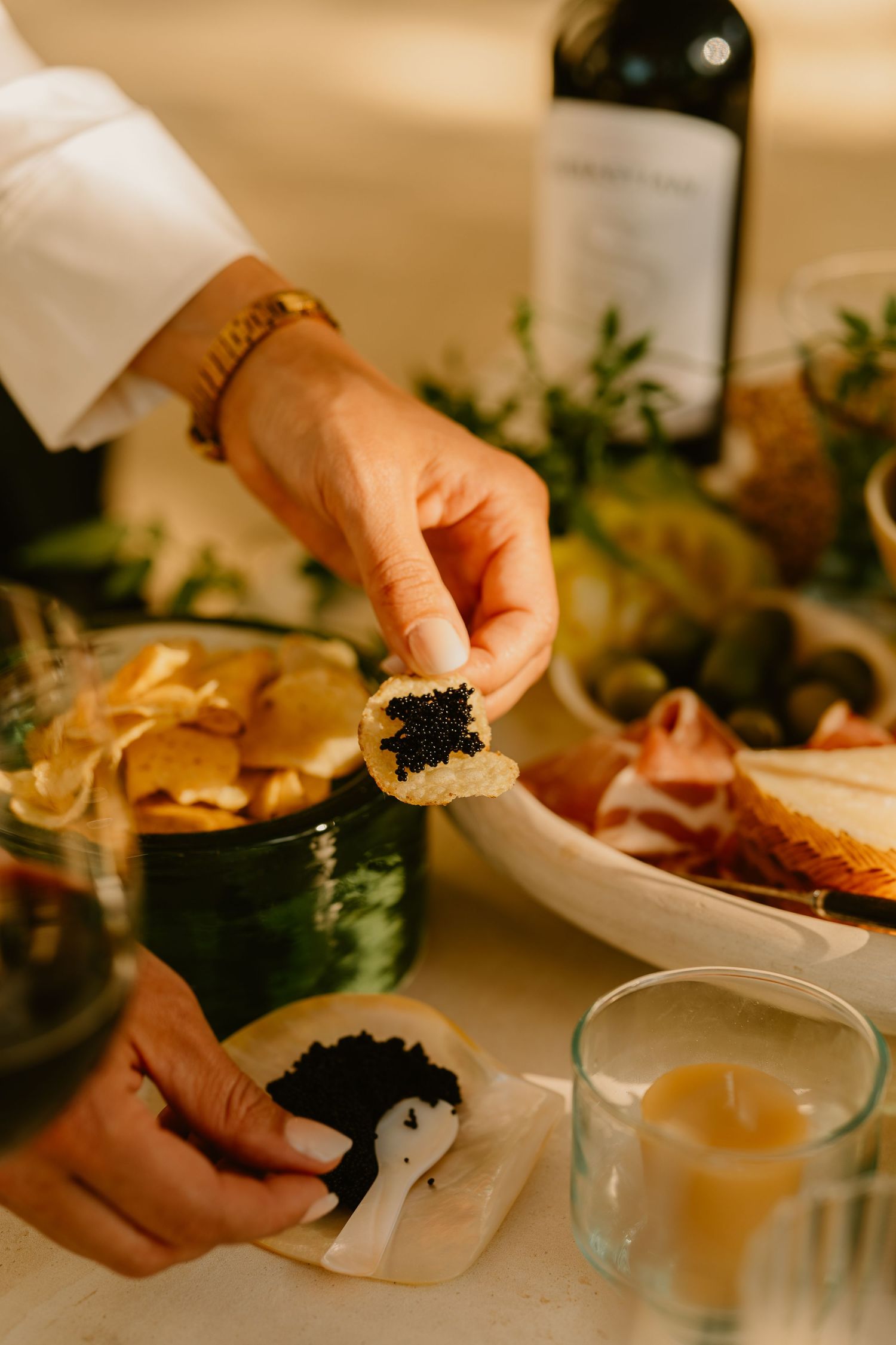 Elegant hand holding caviar-topped cracker at gourmet charcuterie spread with wine bottle and candle.