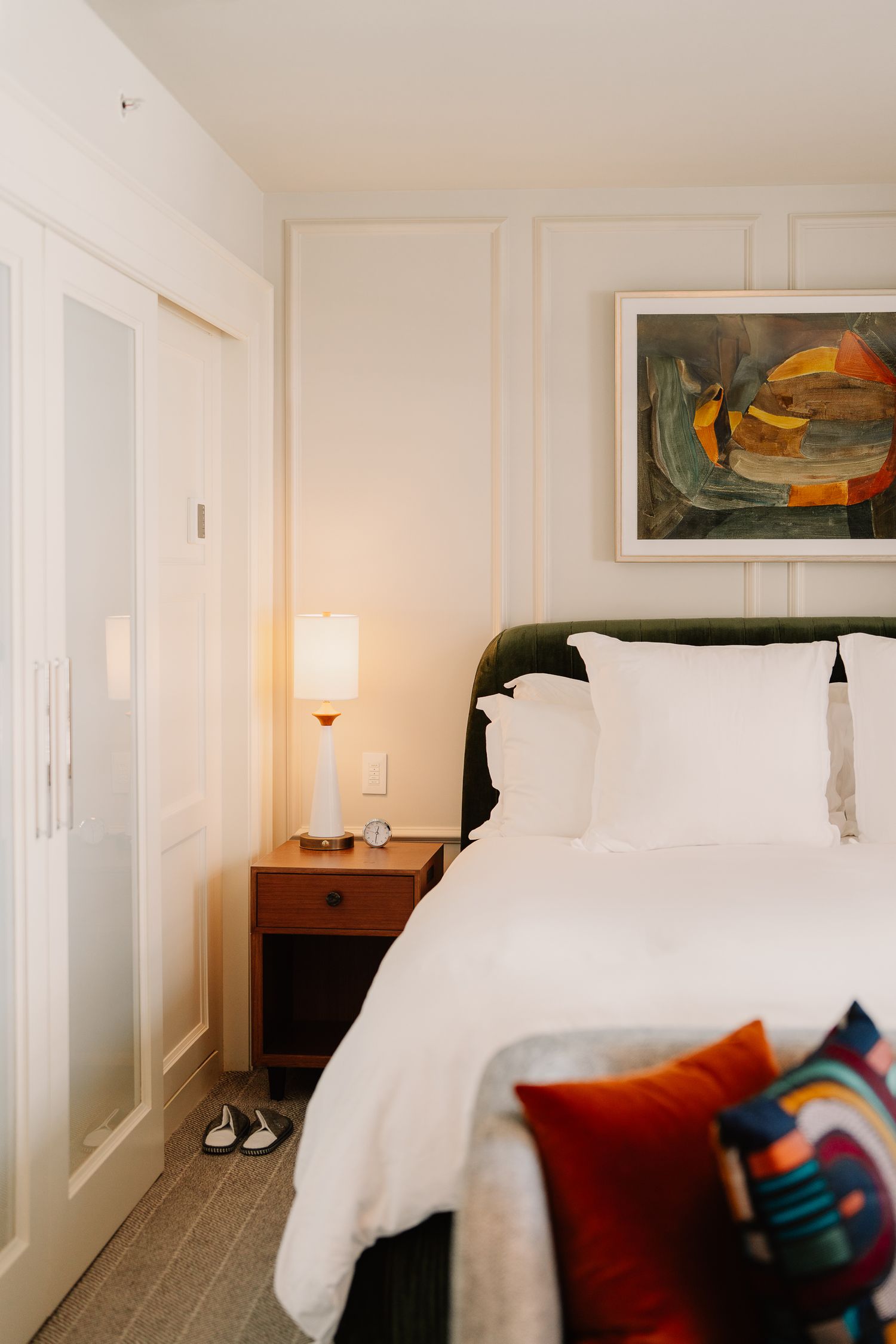 A cozy hotel bedroom with white bedding, colorful throw pillows, and modern artwork featuring orange and blue tones.