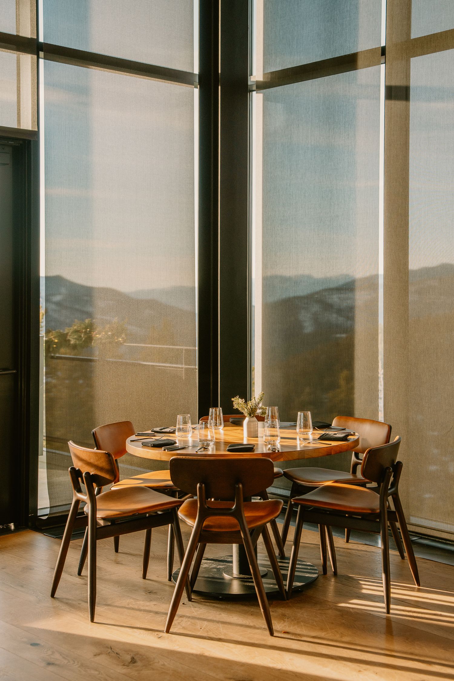A modern dining table with brown leather chairs sits beside floor-to-ceiling windows overlooking mountain views at sunset.