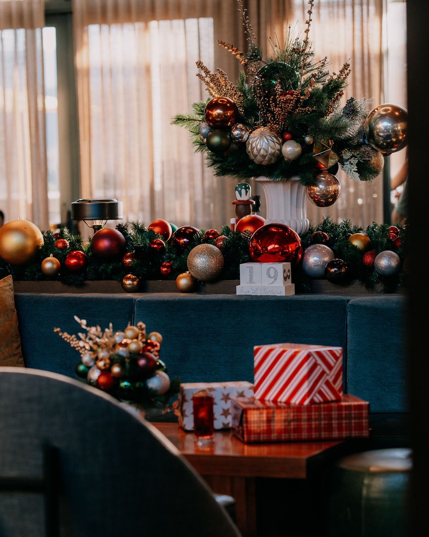 A Christmas scene with red and silver ornaments, wrapped gifts, and evergreen branches on dark tables near curtained windows.